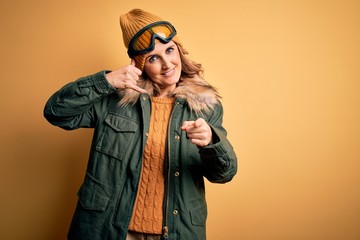 Middle age beautiful blonde skier woman wearing snow sportwear and ski goggles smiling doing talking on the telephone gesture and pointing to you. Call me.