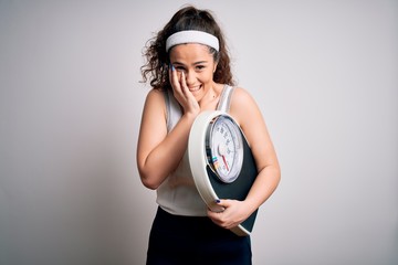 Young beautiful woman with curly hair holding weighing machine over white background laughing and...