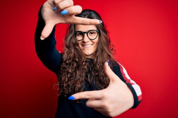 Naklejka premium Young beautiful woman with curly hair wearing sweater and glasses over red background smiling making frame with hands and fingers with happy face. Creativity and photography concept.