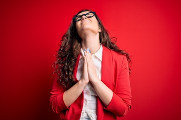 Young beautiful woman with curly hair wearing jacket and glasses over red background begging and...