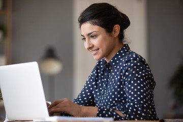 Smiling Indian young woman working on laptop, looking at screen, happy beautiful girl chatting or...