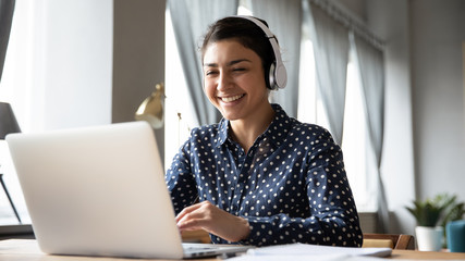 Smiling Indian girl wearing headphones using laptop, looking at screen, happy young female...