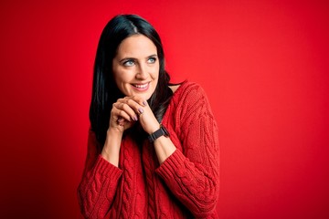 Young brunette woman with blue eyes wearing casual sweater over isolated red background laughing nervous and excited with hands on chin looking to the side