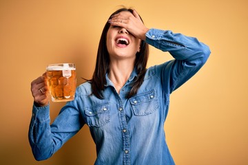 Young woman with blue eyes drinking jar of beer standing over isolated yellow background smiling and laughing with hand on face covering eyes for surprise. Blind concept.