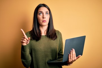 Young brunette woman with blue eyes working using computer laptop over yellow background Pointing...