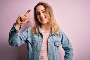 Young beautiful blonde woman wearing casual denim jacket standing over pink background smiling and confident gesturing with hand doing small size sign with fingers looking and the camera. Measure