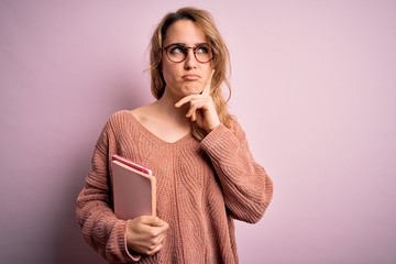 Young beautiful blonde woman wearing glasses holding notebook over pink background serious face...