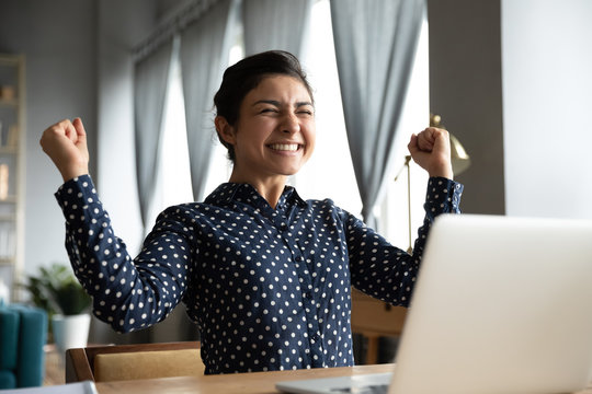 Smiling Indian woman excited by good news, work success, received great shopping or job offer, successful exam results, happy girl showing yes gesture, celebrating win, sitting at desk with laptop