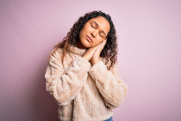 Young beautiful woman with curly hair wearing casual sweater standing over pink background sleeping tired dreaming and posing with hands together while smiling with closed eyes.