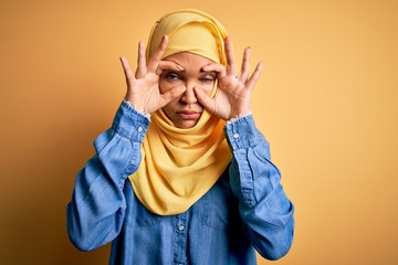 Young beautiful woman with curly hair wearing arab traditional hijab over yellow background Trying to open eyes with fingers, sleepy and tired for morning fatigue