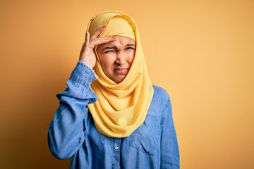 Young beautiful woman with curly hair wearing arab traditional hijab over yellow background worried and stressed about a problem with hand on forehead, nervous and anxious for crisis