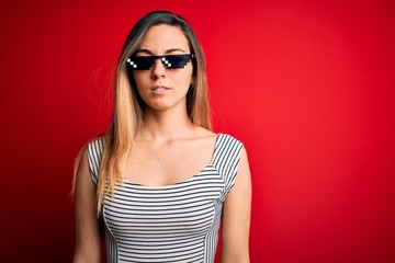 Young beautiful brunette woman wearing funny thug life sunglasses over red background with serious expression on face. Simple and natural looking at the camera.