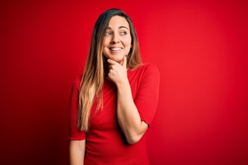 Young beautiful blonde woman with blue eyes wearing casual t-shirt over red background with hand on chin thinking about question, pensive expression. Smiling with thoughtful face. Doubt concept.