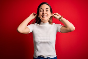 Beautiful blonde woman with blue eyes wearing casual white t-shirt over red background Smiling pulling ears with fingers, funny gesture. Audition problem