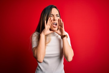 Fototapeta premium Beautiful blonde woman with blue eyes wearing casual white t-shirt over red background Shouting angry out loud with hands over mouth