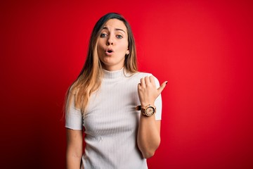 Beautiful blonde woman with blue eyes wearing casual white t-shirt over red background Surprised pointing with hand finger to the side, open mouth amazed expression.