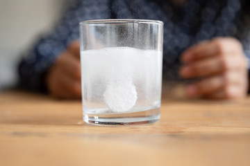 Close up sparkling water glass with dissolving effervescent aspirin pill standing on wooden table,...