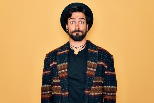 Young handsome hispanic bohemian man wearing hippie style and boho hat puffing cheeks with funny face. Mouth inflated with air, crazy expression.