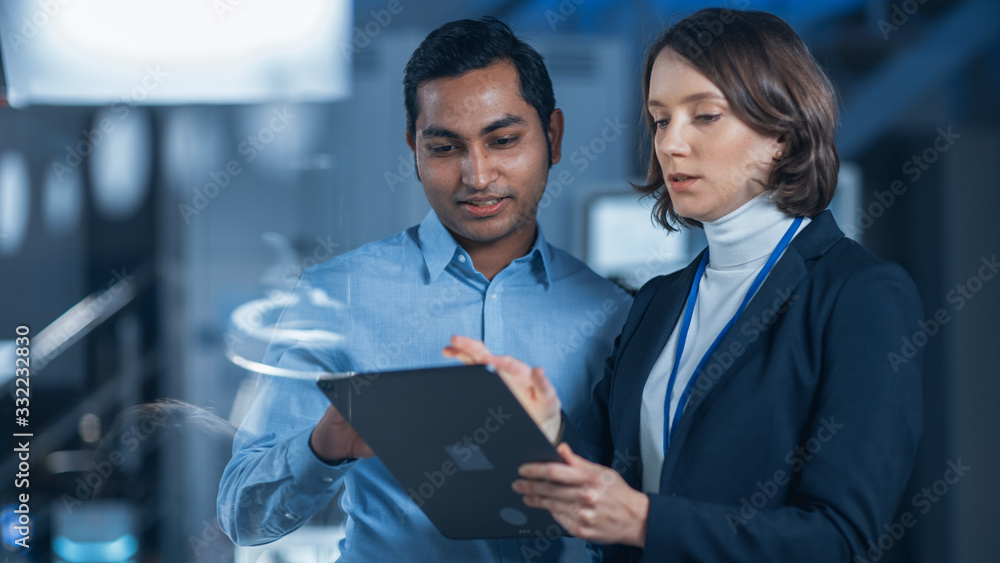 Poster In Technology Research Facility: Female Project Manager Talks With Chief Engineer, they Consult Tablet Computer. Team of Industrial Engineers, Developers Work on Engine Design Using Computers