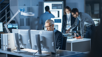 Shot of Industrial Engineer Working in Research Laboratory / Development Center, Using Computer. In the Background Technology Development Laboratory with Scientists, Engineers Working