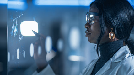 Close-up Shot of an Smart and Beautiful African American Female Scientist Wearing White Coat and Protective Glasses Writes Formula on Glass Whiteboard, References Her Tablet Computer