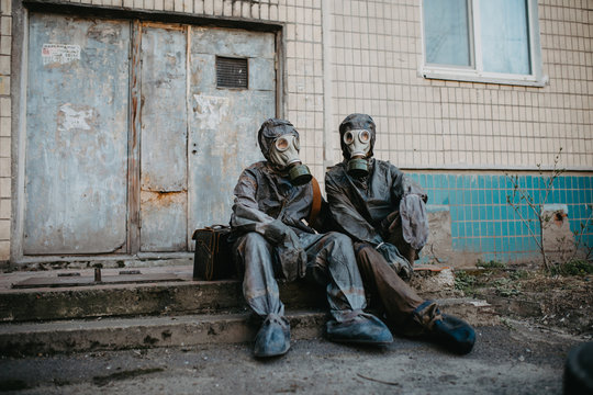 Couple In Love Sits In NBC Protective Suits And Gas Masks Near Building.