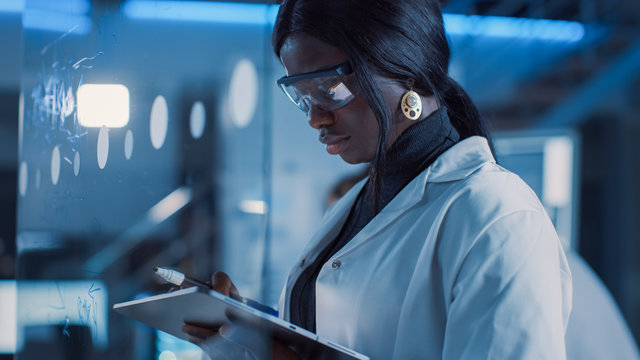In the Research Laboratory Smart and Beautiful African American Female Scientist Wearing White Coat and Protective Glasses Writes Formula on Glass Whiteboard, References Her Tablet Computer