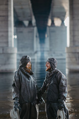 Couple in love holds hands and stands under bridge in NBC protective suits and gas masks in their hands.