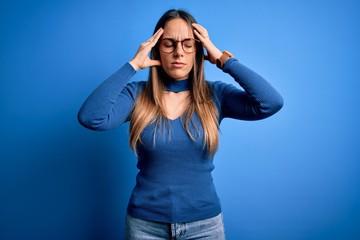 Young beautiful blonde woman with blue eyes wearing glasses standing over blue background with hand on head for pain in head because stress. Suffering migraine.