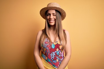 Young beautiful blonde woman wearing swimsuit and summer hat over yellow background winking looking...
