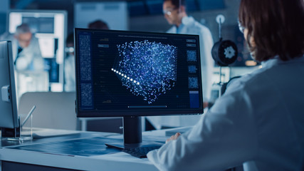 Over the Shoulder Shot: Female IT Scientist Uses Computer Showing System Monitoring and Controlling Program. In the Background Technology Development Laboratory with Scientists, Engineers Working