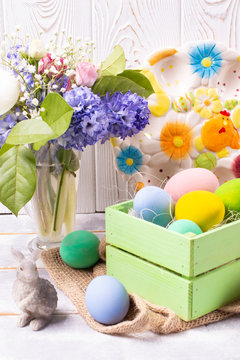 Easter composition with colorful eggs in a wooden crate, a rabbit figurine and a spring bouquet in a vase and an Easter ceramic plate on a white wooden table