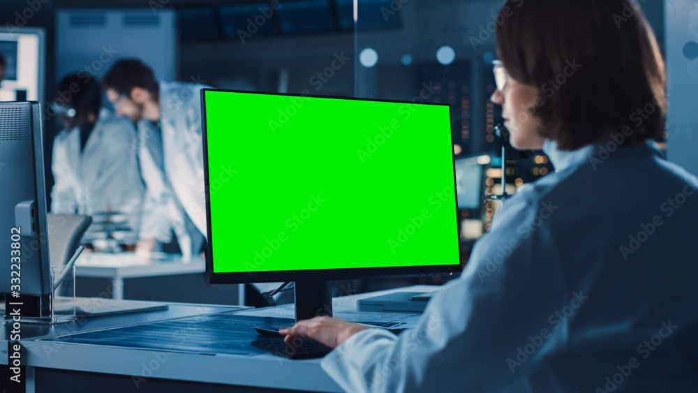 Poster Over the Shoulder Shot: Female IT Scientist Uses Green Mock-up Screen Computer. In the Background Technology Research And Development Laboratory with Scientists, Engineers Working on High Tech Design
