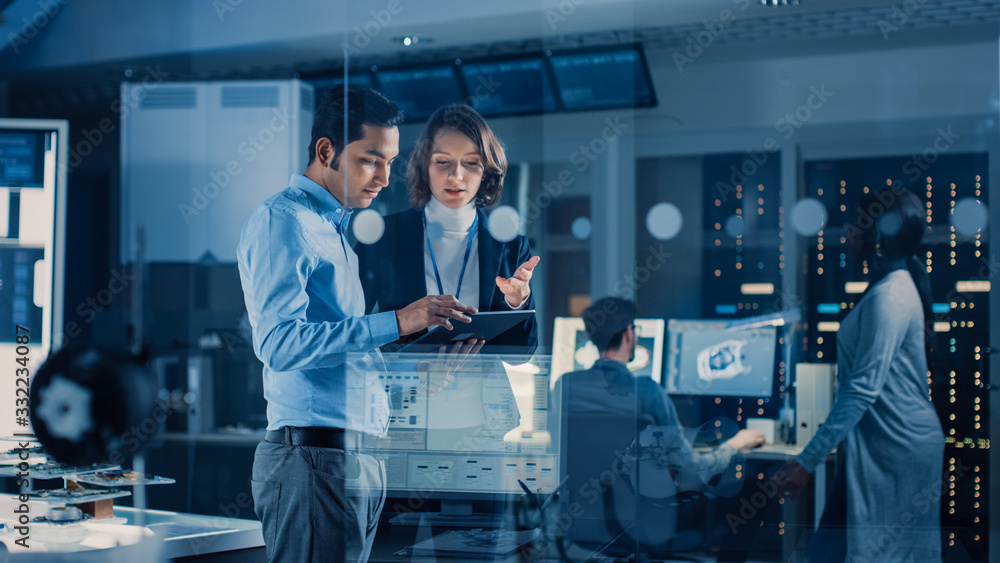 Poster In Technology Research Facility: Female Project Manager Talks With Chief Engineer, they Consult Tablet Computer. Team of Industrial Engineers, Developers Work on Engine Design Using Computers