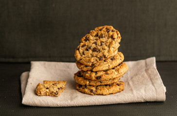 Stack of chocolate chips cookies, biscuits, shortbread on grey napkin. Black background. One cookie bitten. Close up. Tasty crunchy dessert, breakfast, snack. Bakery, coffee house, homemade, recipe