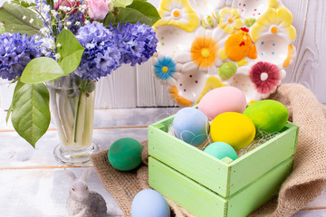 Easter composition with colorful eggs in a wooden crate, a rabbit figurine and a spring bouquet in a vase and an Easter ceramic plate on a white wooden table