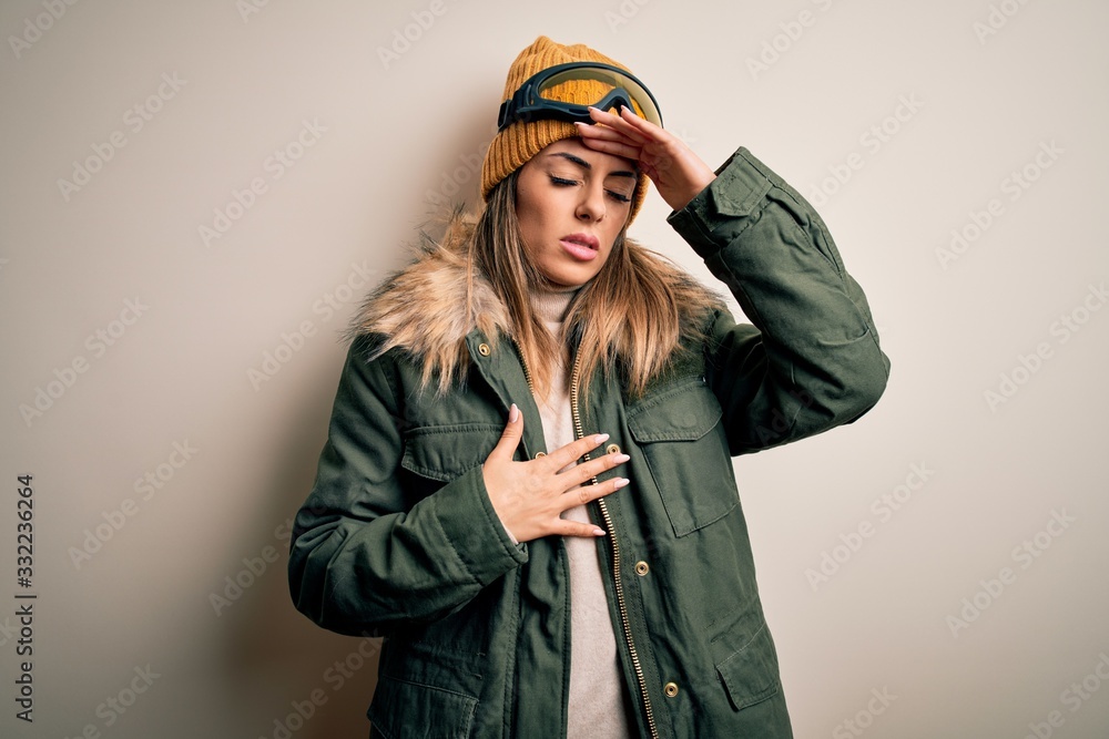 Sticker young brunette skier woman wearing snow clothes and ski goggles over white background touching foreh