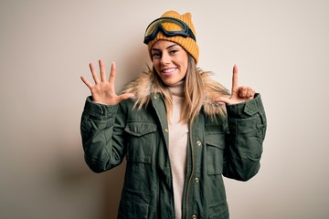 Young brunette skier woman wearing snow clothes and ski goggles over white background showing and pointing up with fingers number seven while smiling confident and happy.