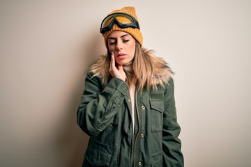 Young brunette skier woman wearing snow clothes and ski goggles over white background touching mouth with hand with painful expression because of toothache or dental illness on teeth. Dentist