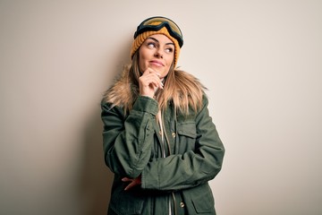 Young brunette skier woman wearing snow clothes and ski goggles over white background with hand on chin thinking about question, pensive expression. Smiling with thoughtful face. Doubt concept.