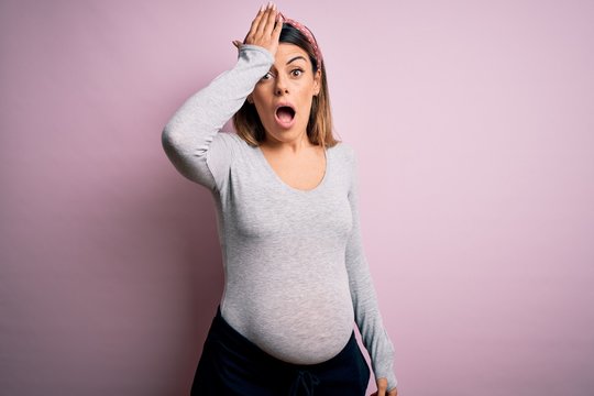 Young beautiful brunette woman pregnant expecting baby over isolated pink background surprised with hand on head for mistake, remember error. Forgot, bad memory concept.