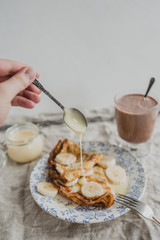 A hand is pouring condensed milk on crepe. Pancakes with bananas, condensed milk and cocoa. Tasty breakfast.