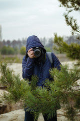 A female photographer with a professional camera peeks out from behind tree branches. Professional photographer. Journalist. Hunting.