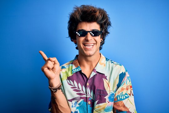 Young handsome man wearing thug life glasses standing over isolated blue background with a big smile on face, pointing with hand and finger to the side looking at the camera.