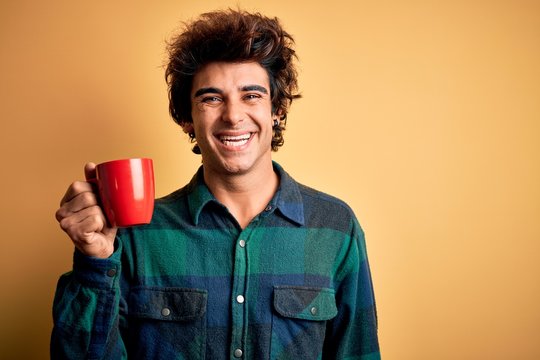 Young handsome man drinking cup of coffee standing over isolated yellow background with a happy face standing and smiling with a confident smile showing teeth