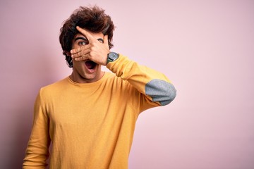 Young handsome man wearing yellow casual t-shirt standing over isolated pink background peeking in shock covering face and eyes with hand, looking through fingers with embarrassed expression.
