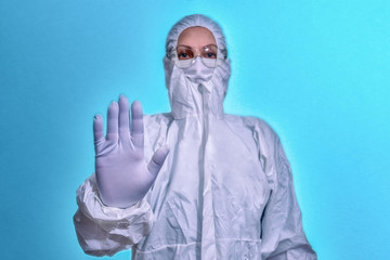 a woman in overalls, safety glasses, a protective mask and white gloves raises her hand in a Stop...