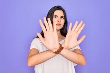 Young beautiful brunette woman wearing casual white t-shirt over purple background Rejection expression crossing arms doing negative sign, angry face