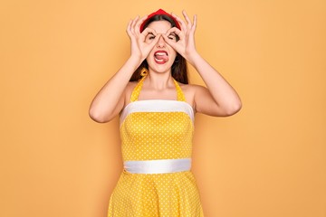 Young beautiful pin up woman wearing 50s fashion vintage dress over yellow background doing ok...