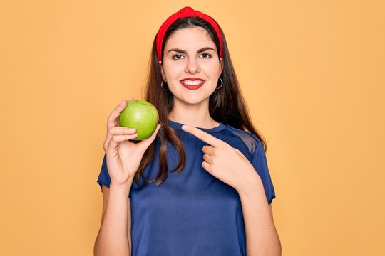 Young beautiful girl eating fresh organic healthy green apple over yellow background very happy pointing with hand and finger